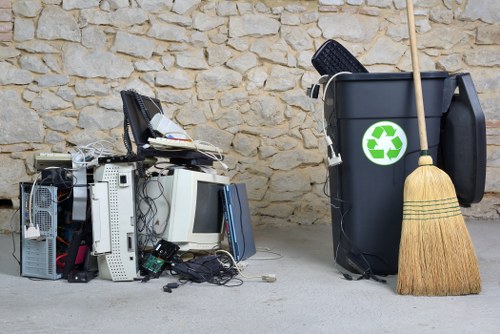 The image displays a collection of discarded electronic waste, including old computer towers, monitors, and various cables, situated on a paved surface against a stone wall background. To the right of the electronic waste, there is a large black waste bin with a green recycling symbol on its front, positioned next to a natural straw broom with a wooden handle leaning against it. The items are arranged outdoors, possibly on a driveway or service area, and appear to be awaiting rubbish removal or recycling collection. The scene underscores professional waste management services provided by Commercial Waste Thornton Heath for disposing of electronic and general rubbish responsibly within the local postcode area.