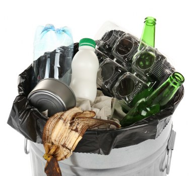 A metal waste bin lined with a black plastic bag, filled with mixed rubbish including clear plastic bottles with blue caps, a white plastic bottle with a green cap, a crumpled rusty metal can, broken black plastic items such as camera casings, and a piece of dried, possibly wooden or cardboard material. The waste is piled towards the top of the bin, with some items overlapping and spilling slightly over the edge of the bag. The background is plain white, emphasizing the contents and the metallic surface of the bin, which appears to be situated indoors or in a controlled environment. The collection of items reflects common household or commercial waste, suitable for rubbish removal services in Thornton Heath or nearby areas, contributing to waste management and disposal solutions.