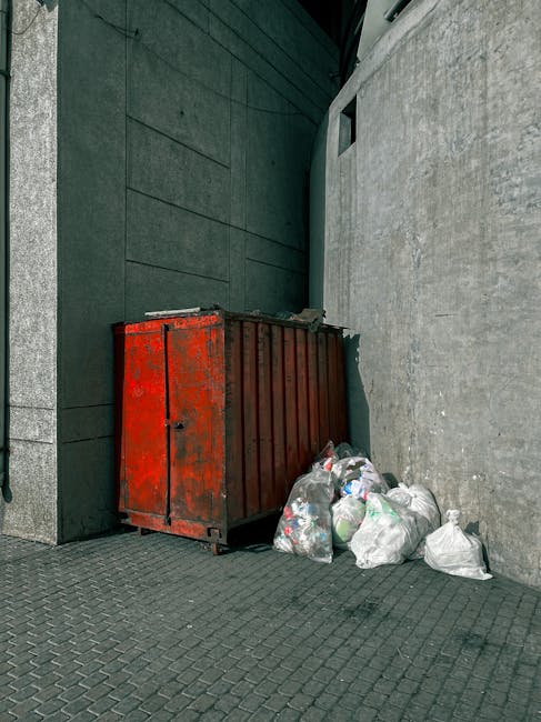 A commercial waste collection truck parked on a narrow urban street with cobblestone pavement, actively engaged in rubbish removal. The vehicle is predominantly white with some rusted metal surfaces v